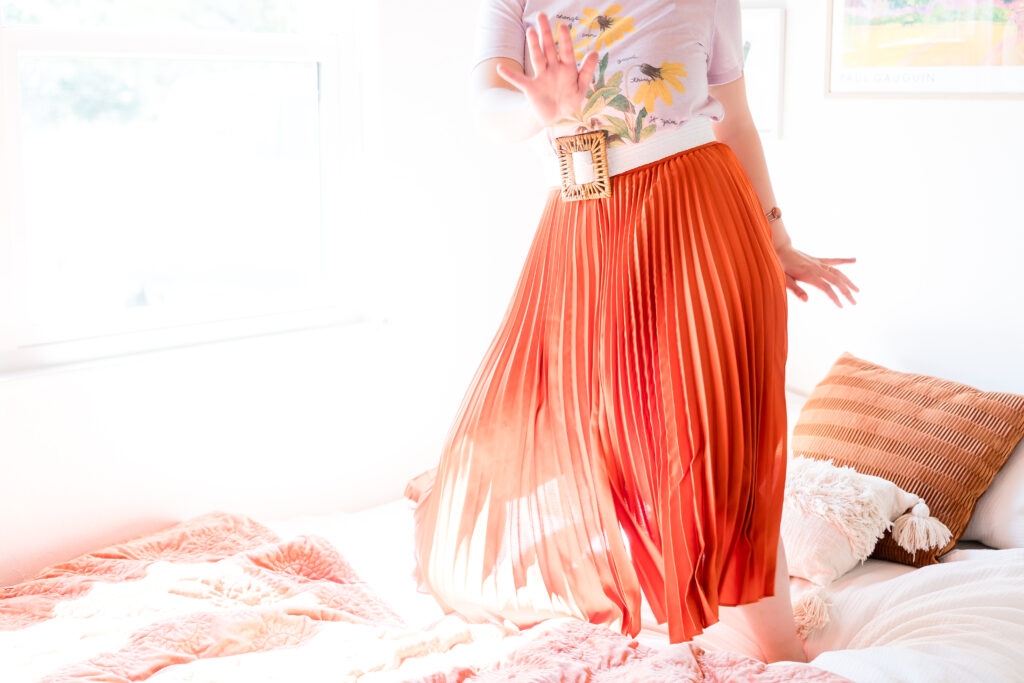 A woman wearing an orange skirt, jumping up and down on a bed as the sunlight leaks in to an Austin, Texas bedroom.
