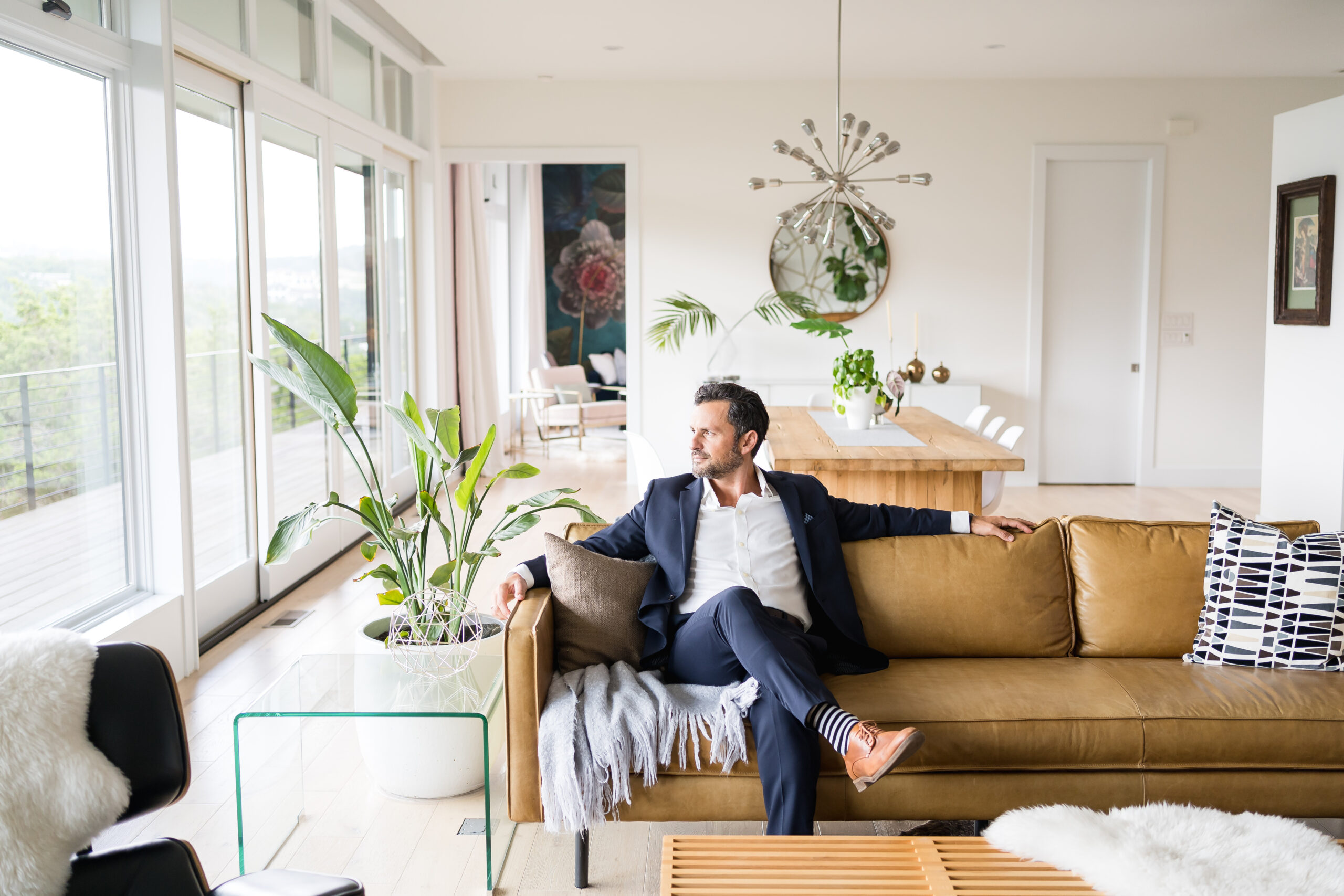 Man in a blue suit, sitting on a brown leather couch in an Austin home, posing for new headshot photos with Alissa Cordoba Photography.