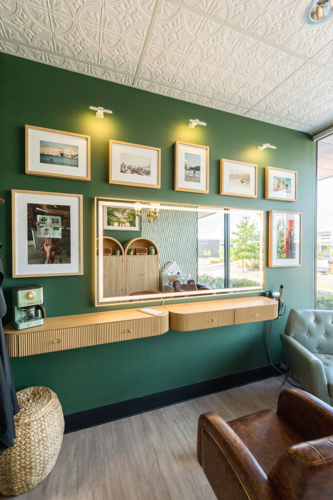 Wide daytime shot of the full green private hair salon suite at Verano Luxe in Leander near Austin Texas, featuring gallery wall, lighted vanity mirror, and leather seating